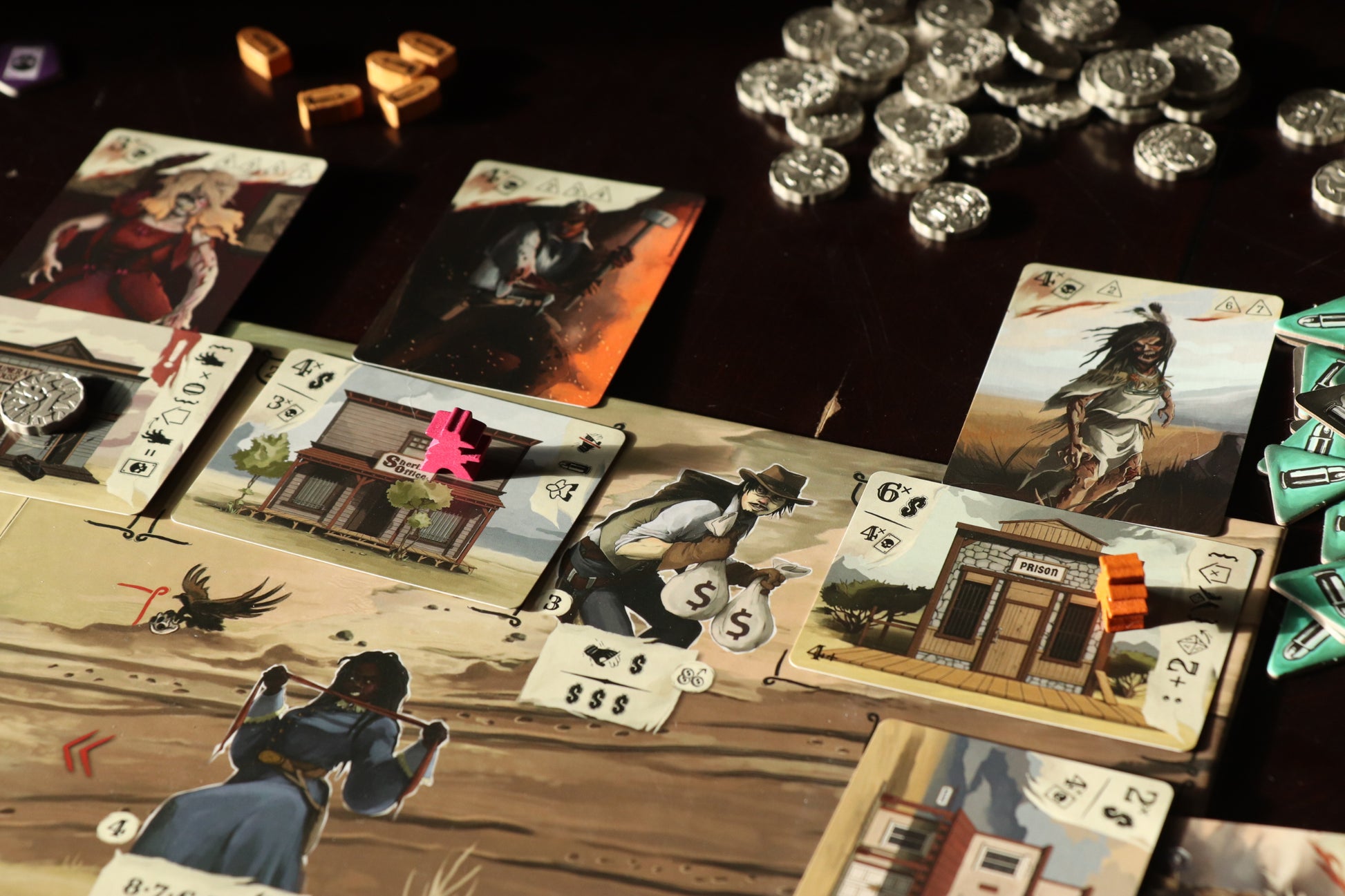 Close-up of a Western-themed board game, cards show characters, buildings, with pink and orange meeples, and silver coins