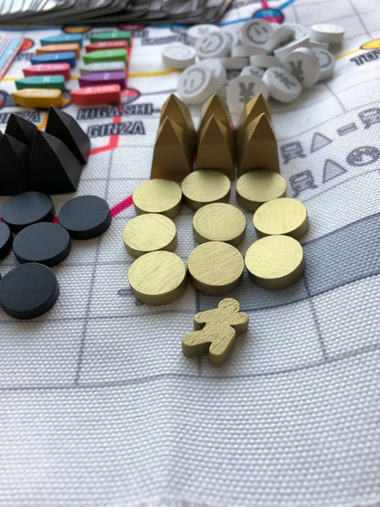 Various board game pieces, including colourful rectangular tokens, gold and black discs, gold pyramids, a gold meeple, and white currency tokens, are arranged on a white grid-patterned game board