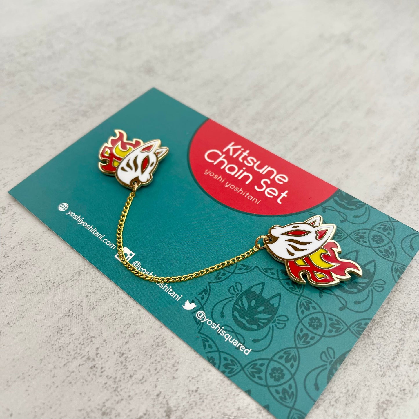 Two white, red, yellow Kitsune fox mask enamel pins connected by a gold chain, displayed on a teal card.