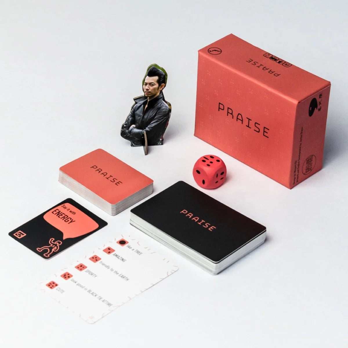 Jordan Draper Games Praise Card Game components displayed: a red box, stacks of red and black cards, a red die, and a cardboard standee of a man