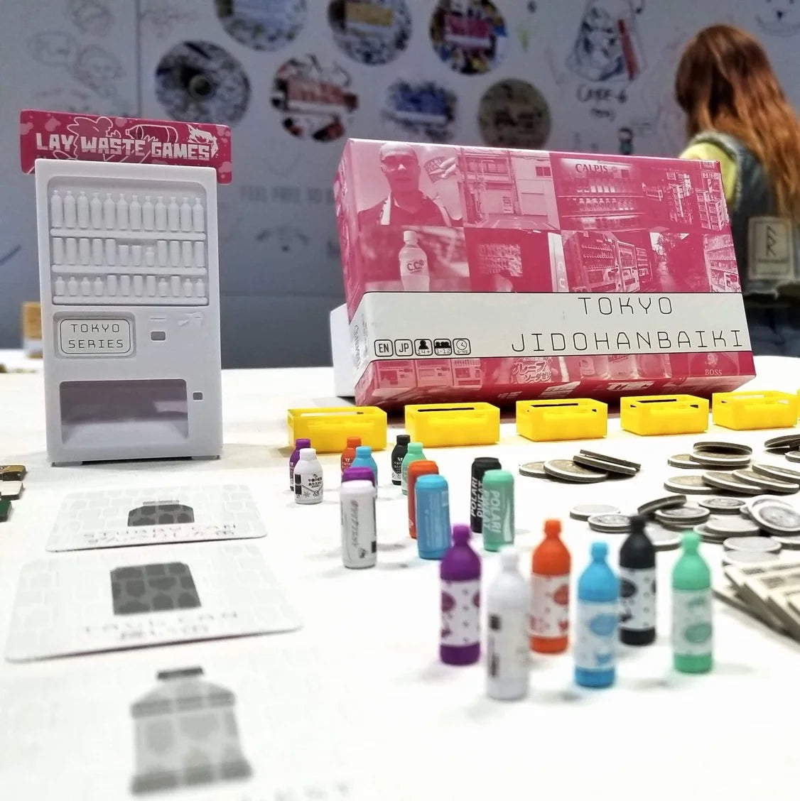 The Tokyo Jidohanbaiki board game features a miniature white vending machine, colourful bottles, game cards, yellow trays, and stacks of coins on a table