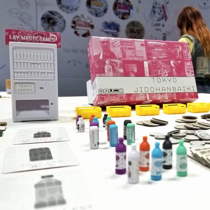The Tokyo Jidohanbaiki board game features a miniature white vending machine, colourful bottles, game cards, yellow trays, and stacks of coins on a table