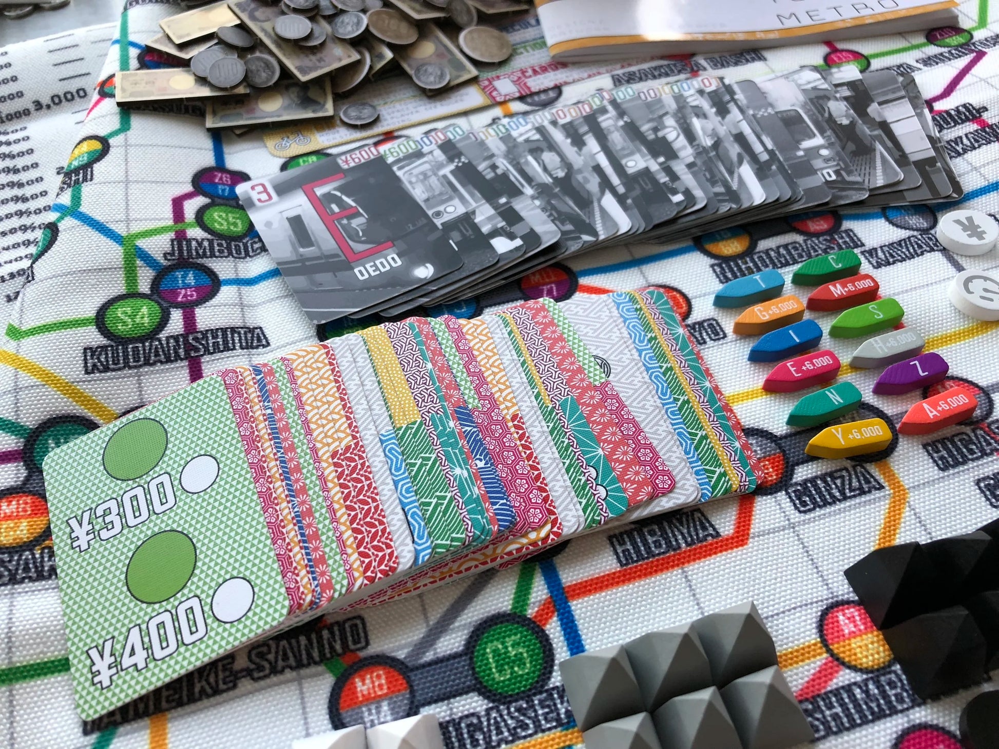 Components of the Tokyo Metro board game are spread on a subway map board, showing train cards, patterned cards, money, and colourful game pieces