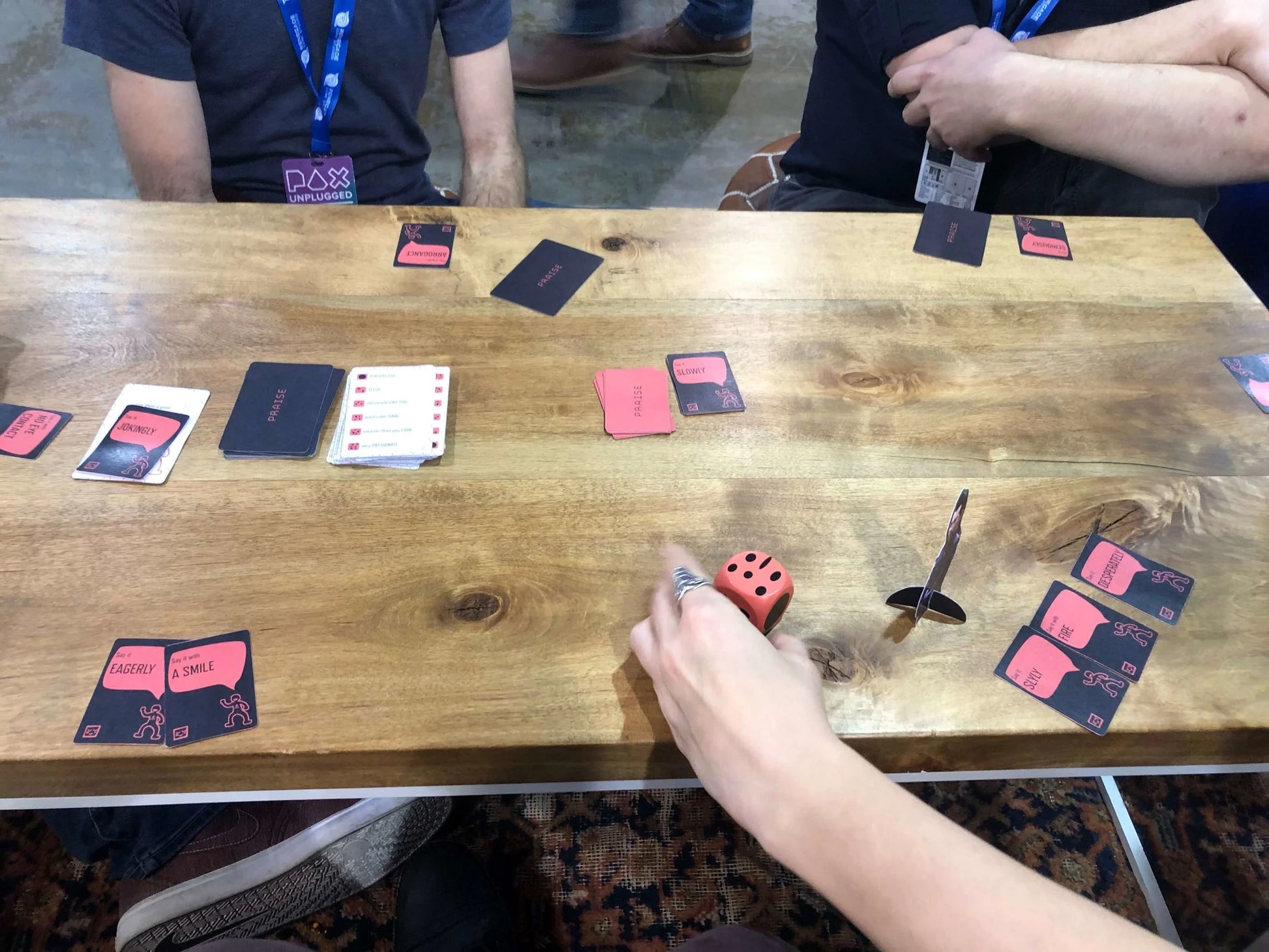 People play the Praise Card Game on a wooden table with various black and red cards, a red die, and a cardboard standee
