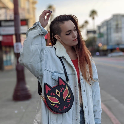 Person with shaved side and long red hair wears a denim jacket and fox mask purse, posing on a city street.