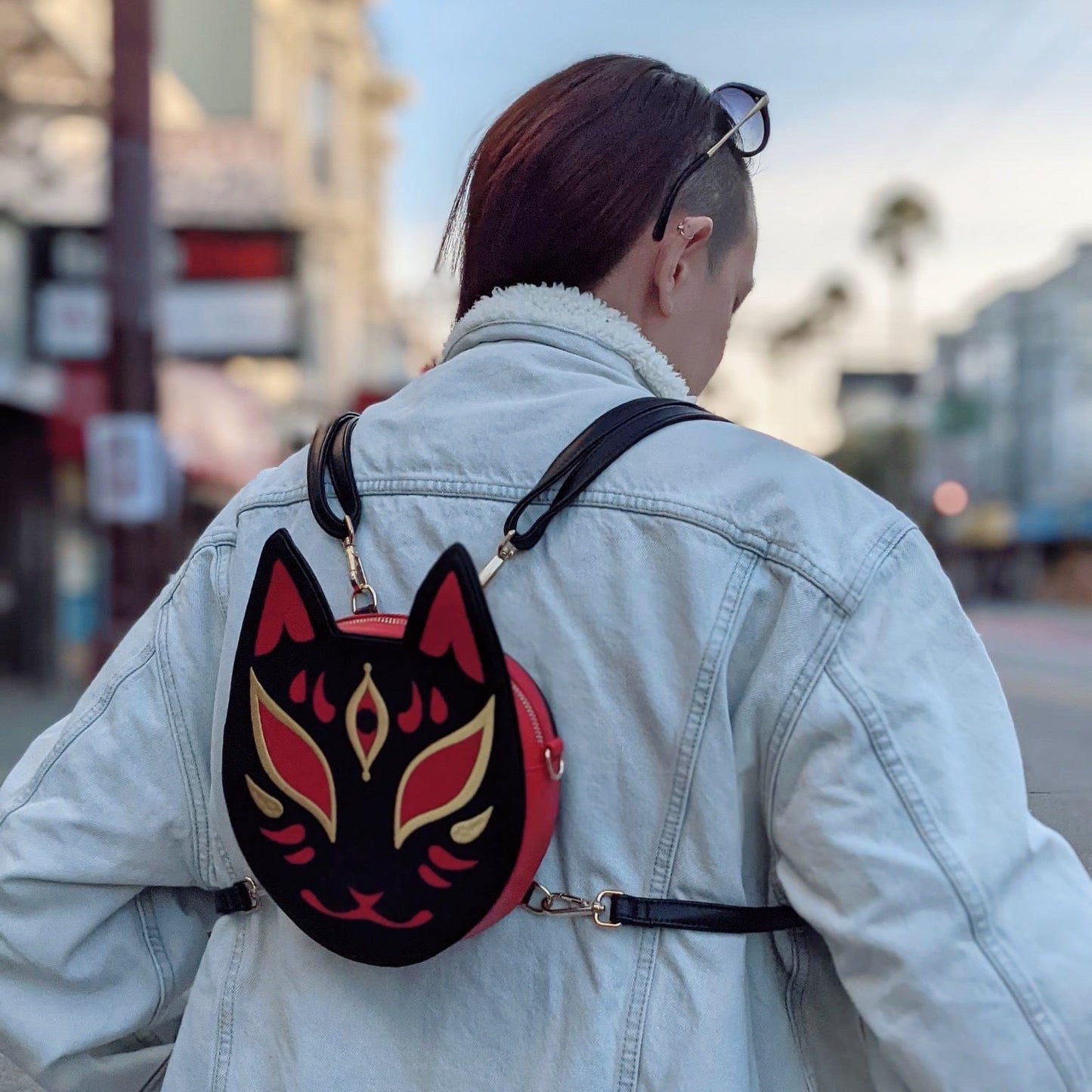 A person in a denim jacket carries a black and red kitsune mask-shaped backpack while walking on an urban street.