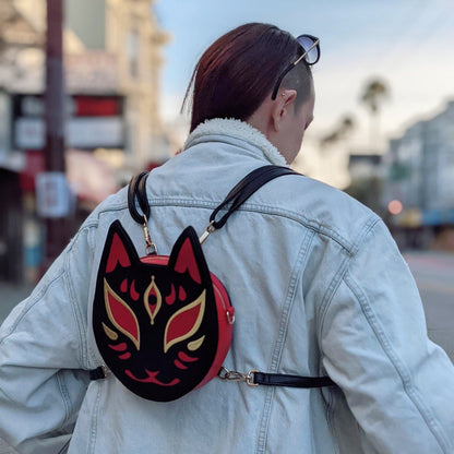 A person in a denim jacket carries a black and red kitsune mask-shaped backpack while walking on an urban street.
