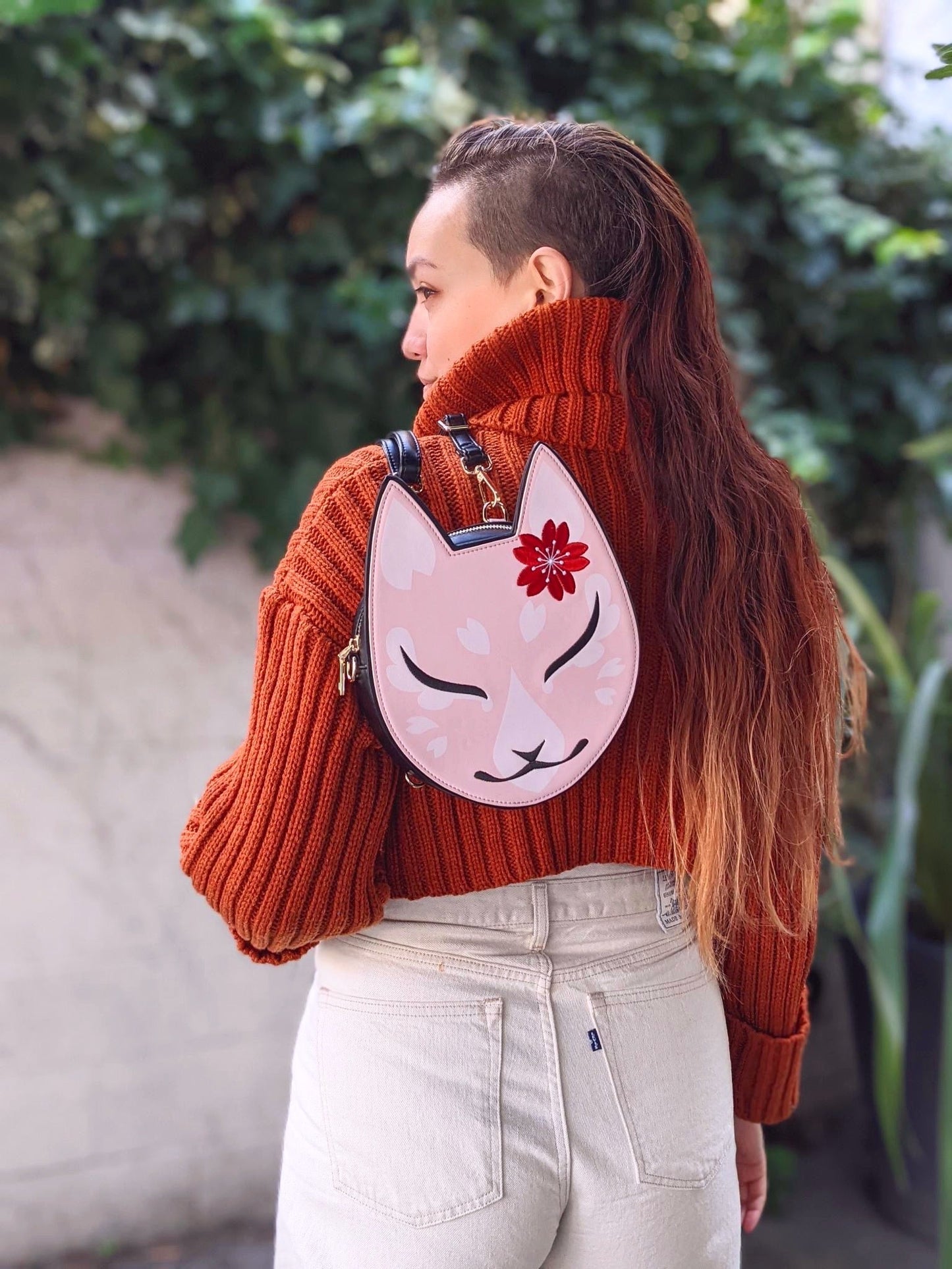 Person from behind in orange sweater and jeans, wearing a pink fox-face backpack with a red flower, looking left.