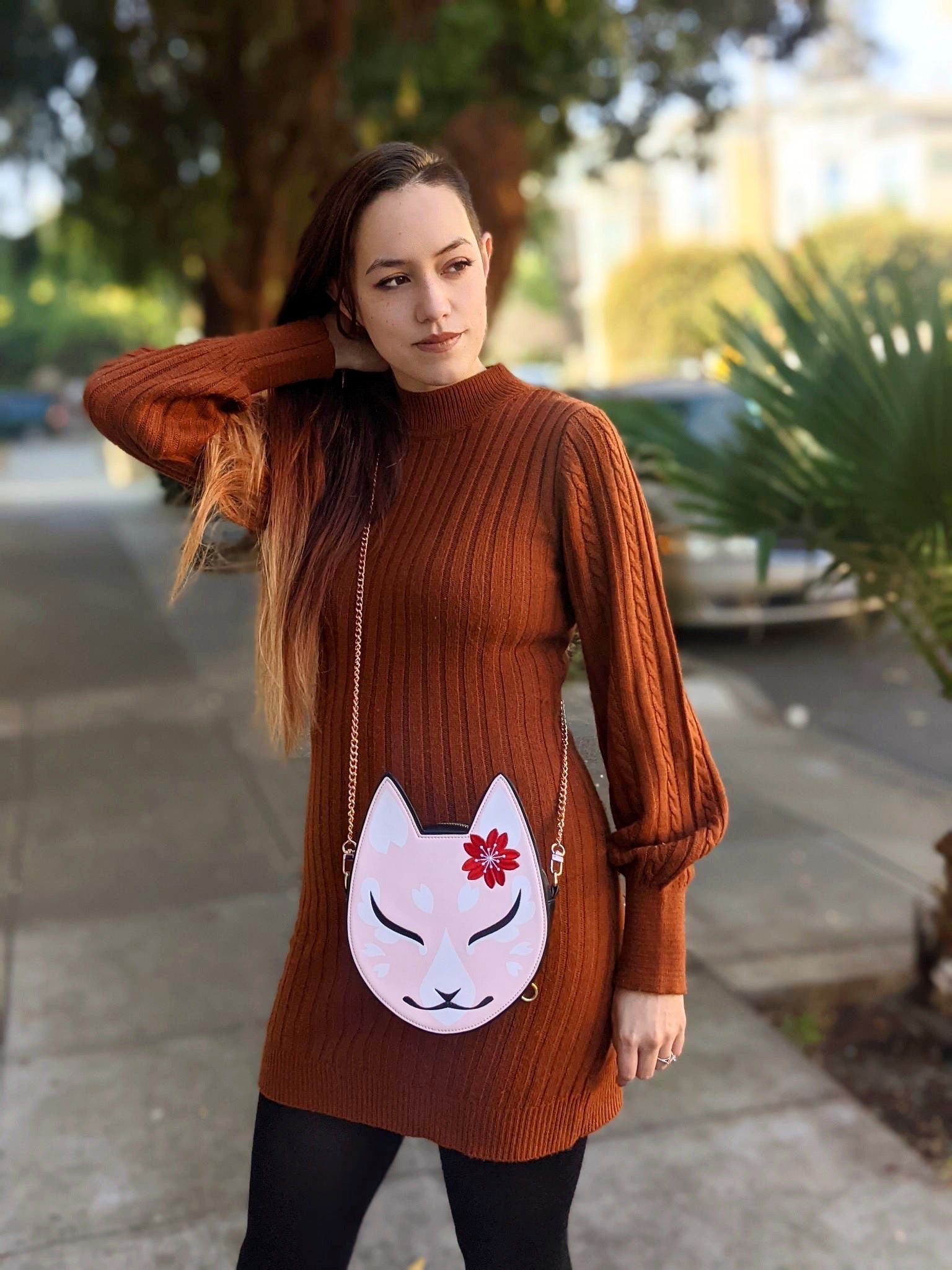 A woman in a rust-colored sweater dress and pink kitsune mask purse, posing with hand in hair on a sunny sidewalk.