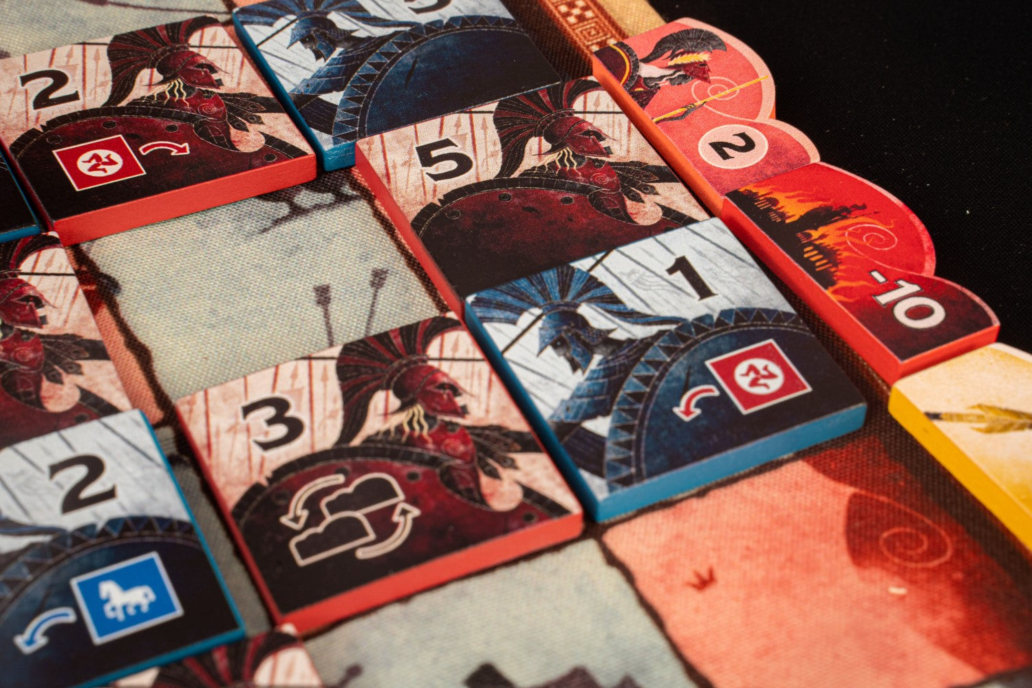 Close-up of a board game with red and blue Spartan warrior tiles, numbered and showing various action symbols, on a game mat