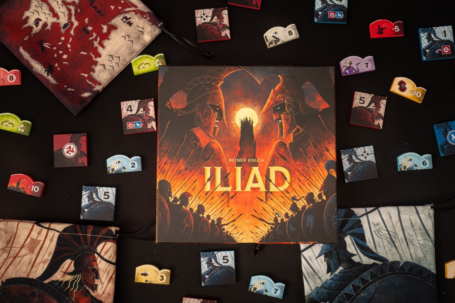 Overhead view of the ILIAD board game box, featuring two helmeted warriors, surrounded by tokens and game bags on a dark surface
