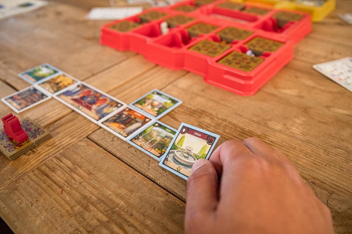 Hand holding a Fountain card from the board game Mariposas near other game pieces on a wooden table