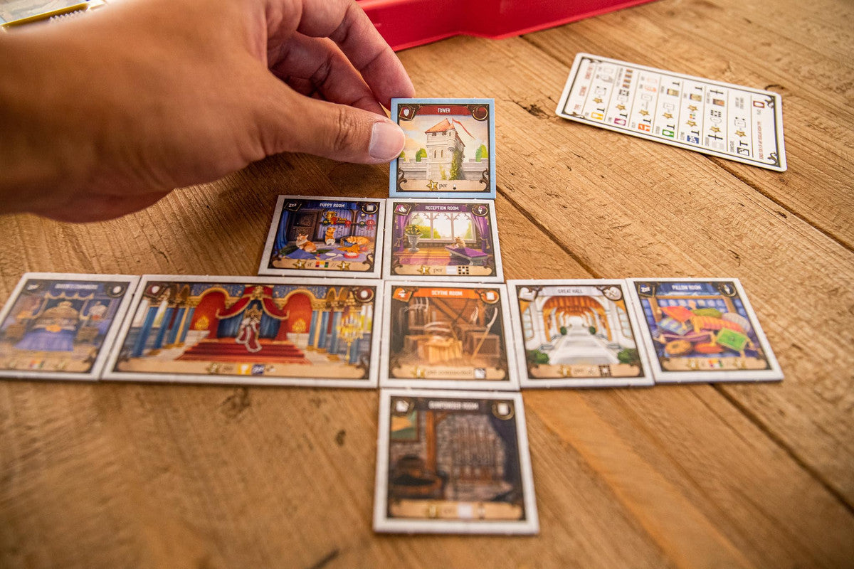 Hand holding a square game tile with a tower building illustration over other game tiles laid out on a wooden table