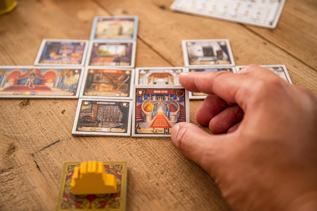 Hand places a Grand Foyer room card on a wooden table with other room cards laid out from the board game Castles of Mad King Ludwig