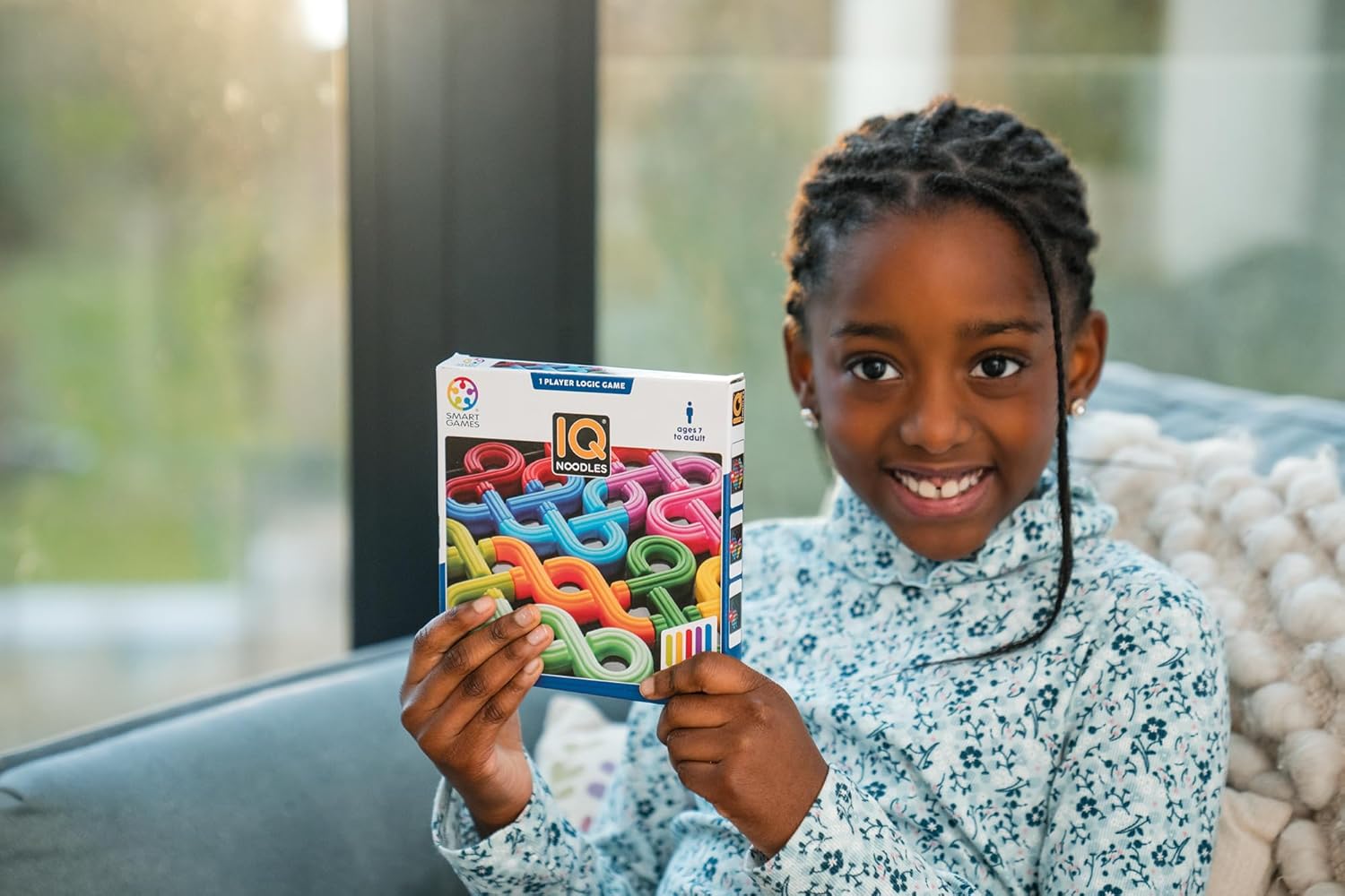 A happy young girl smiles while holding a colorful IQ Noodles logic game box
