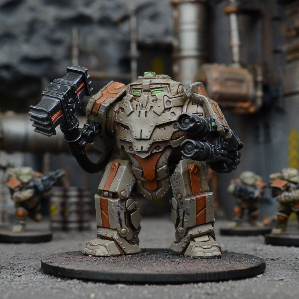 A tabletop miniature of a beige and orange mech with green eyes, wielding a power hammer and arm cannon in an industrial setting.