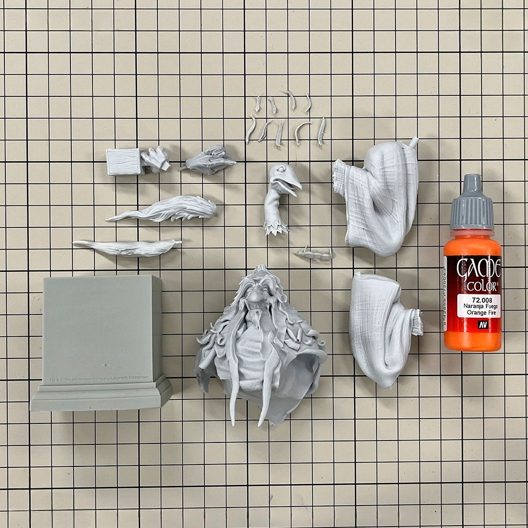 Grey unassembled miniature kit parts: a wizard bust, bird creature, and other pieces, with an orange paint bottle on a grid mat.