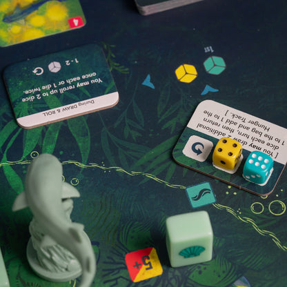 Tabletop game components: dolphin miniature, ability cards, and dice (yellow, teal, mint) on a dark green underwater game mat