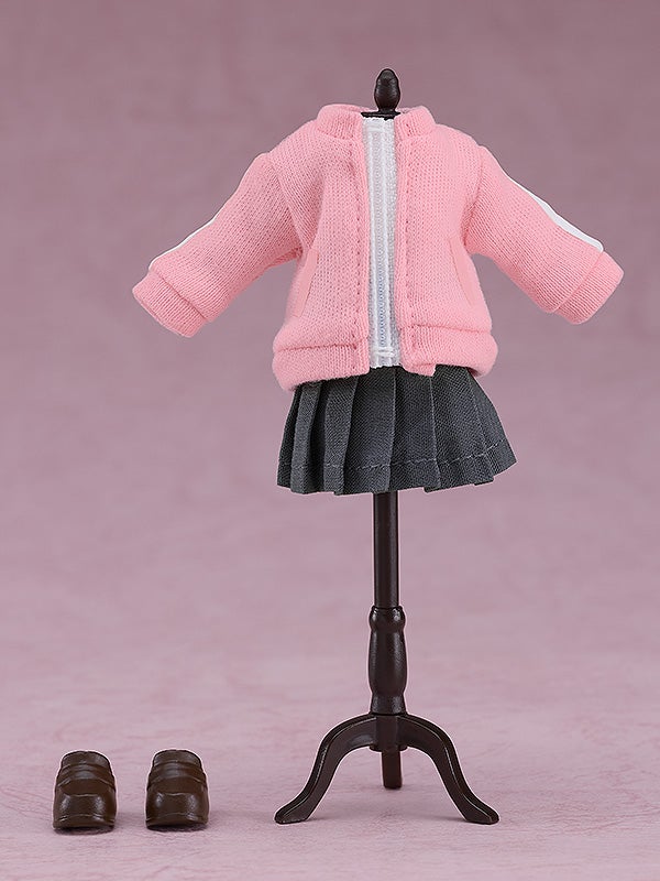 Pink knit track jacket with white stripe detail over a dark gray pleated skirt on a mannequin with miniature brown loafers beside