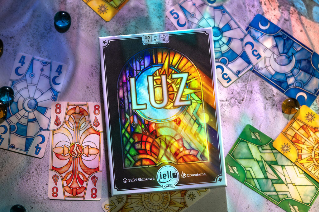 Vibrant 'LUZ' card game box with glowing stained-glass art surrounded by matching cards and scattered beads