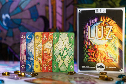 Luz board game box and a row of colorful stained glass-style cards featuring star, moon, sun, and fire symbols. Game tokens are scattered