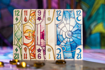 A line of colorful game cards standing upright, each with a stained-glass design, numbers, and elemental symbols. Blurry golden tokens foreground