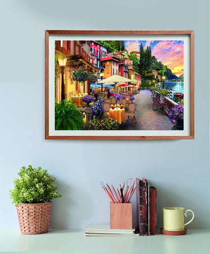 Framed picture of a vibrant waterside town, featuring outdoor cafes, colorful buildings, abundant flowers, a lake, and a sunset. Above a desk.
