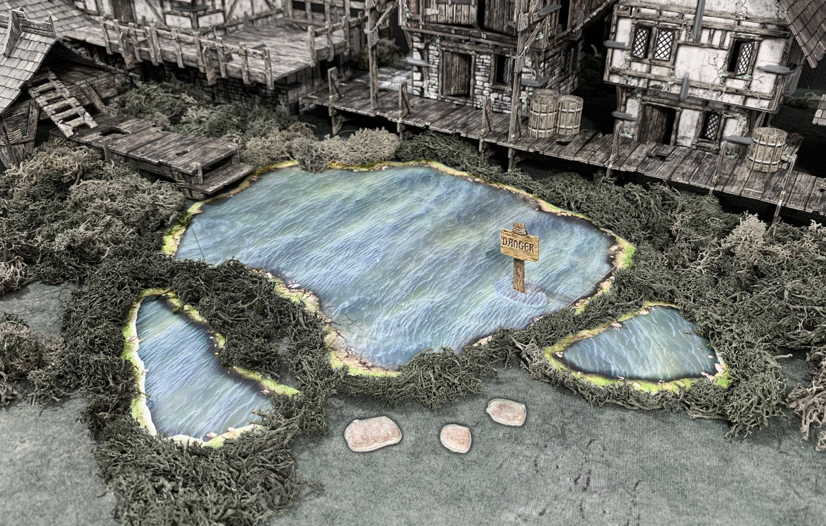 A miniature medieval village terrain: A lake with a &#39;DANGER&#39; sign, two small ponds, and surrounding mossy docks and buildings.