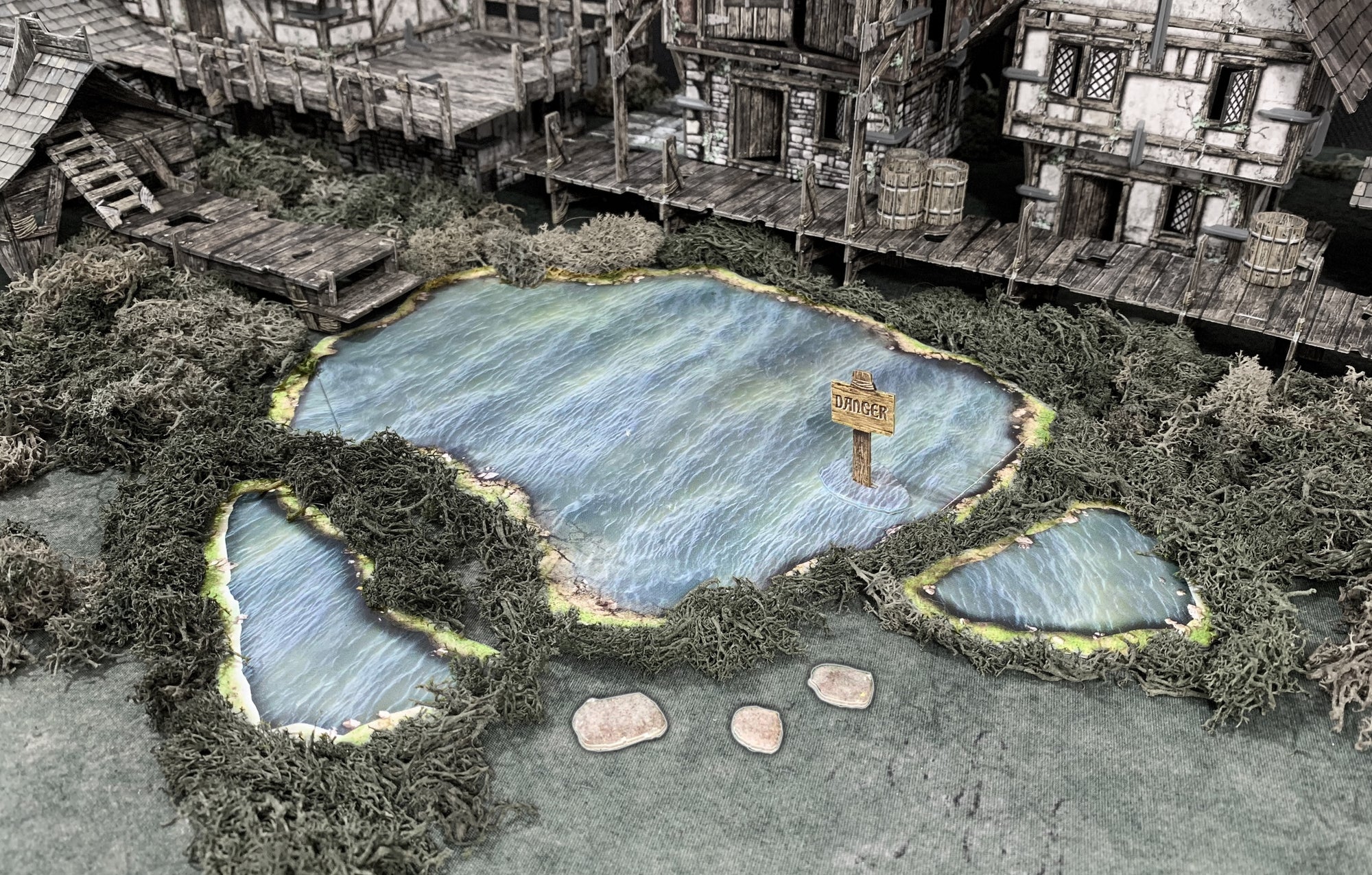 A miniature medieval village terrain: A lake with a 'DANGER' sign, two small ponds, and surrounding mossy docks and buildings.