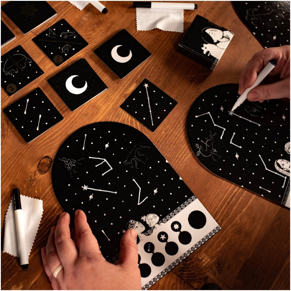 Hands drawing constellations on black game boards with white markers for a celestial-themed game