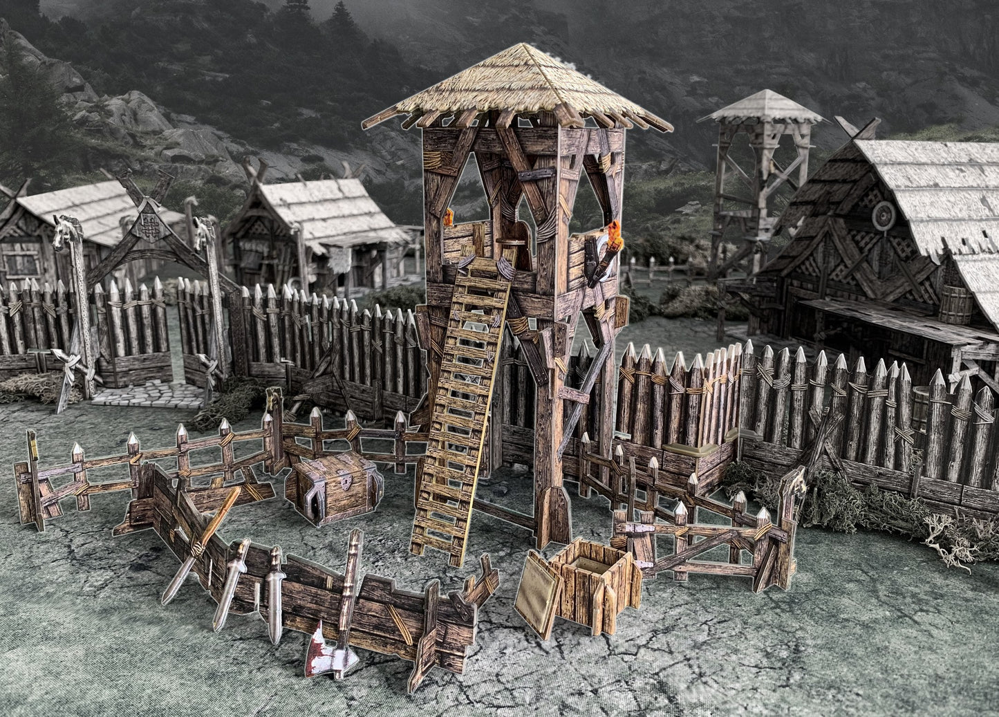 Tabletop Viking fortress model: central watchtower, wooden palisades, thatched houses, and scattered weapons.
