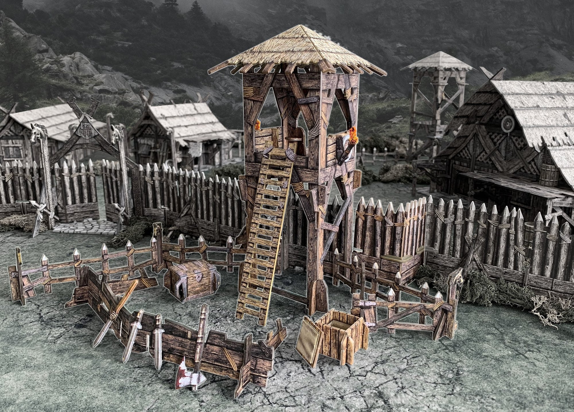 Tabletop Viking fortress model: central watchtower, wooden palisades, thatched houses, and scattered weapons.