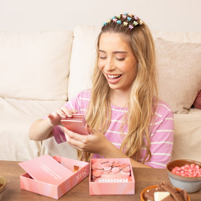 A smiling woman with colorful hair clips holds pink Sync game cards, with open game boxes and snacks on a wooden table.