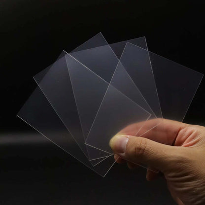 Hand holding fanned-out transparent rectangular plastic sheets, reflecting light, against a dark background