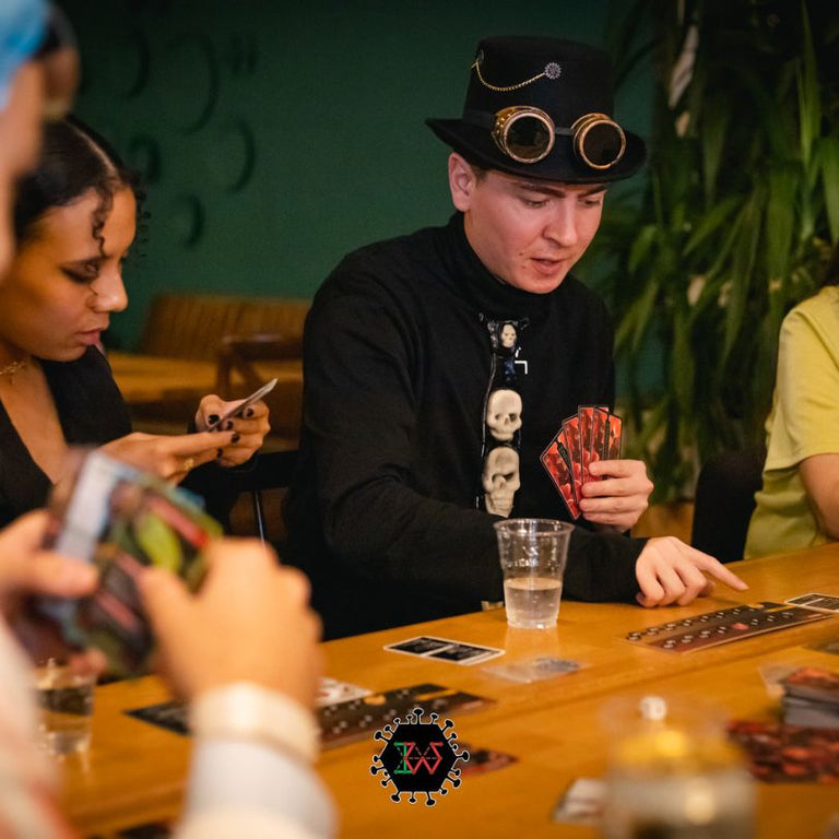 A man in a black top hat and goggles plays a card game, holding red cards, at a table with others