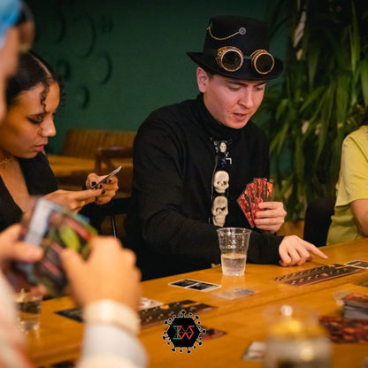 A man in a black top hat and goggles plays a card game, holding red cards, at a table with others