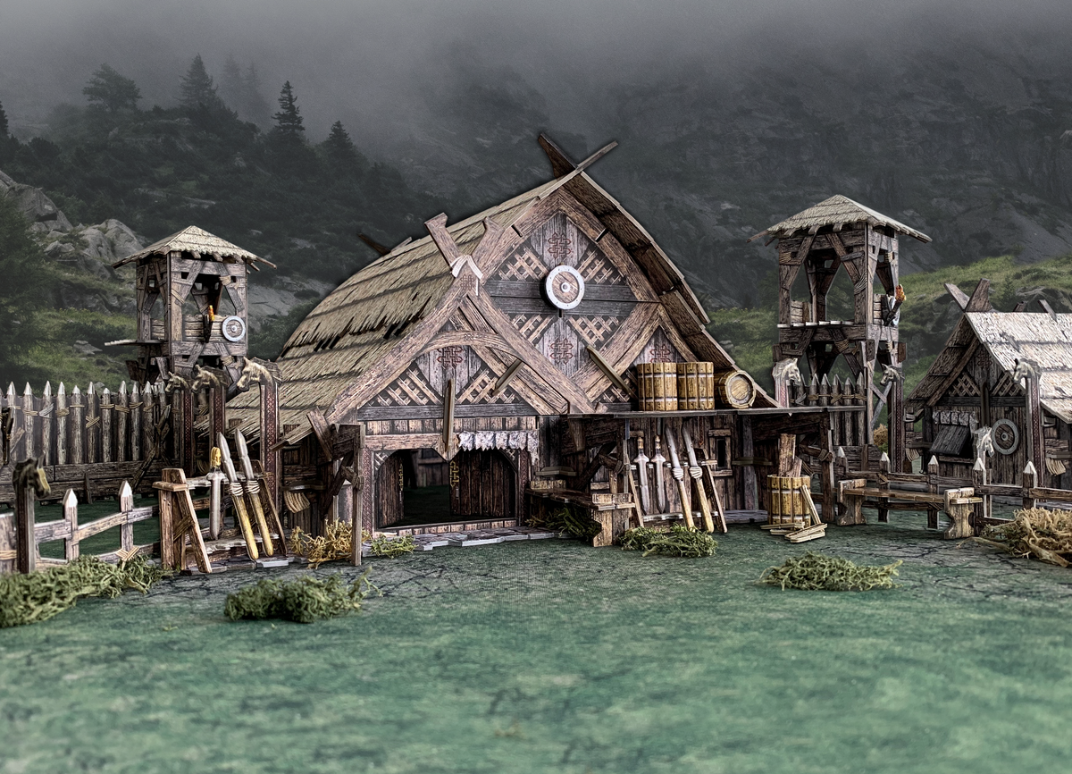 Detailed miniature Viking village: a longhouse, two watchtowers, wooden fence, and weapons with misty mountains.