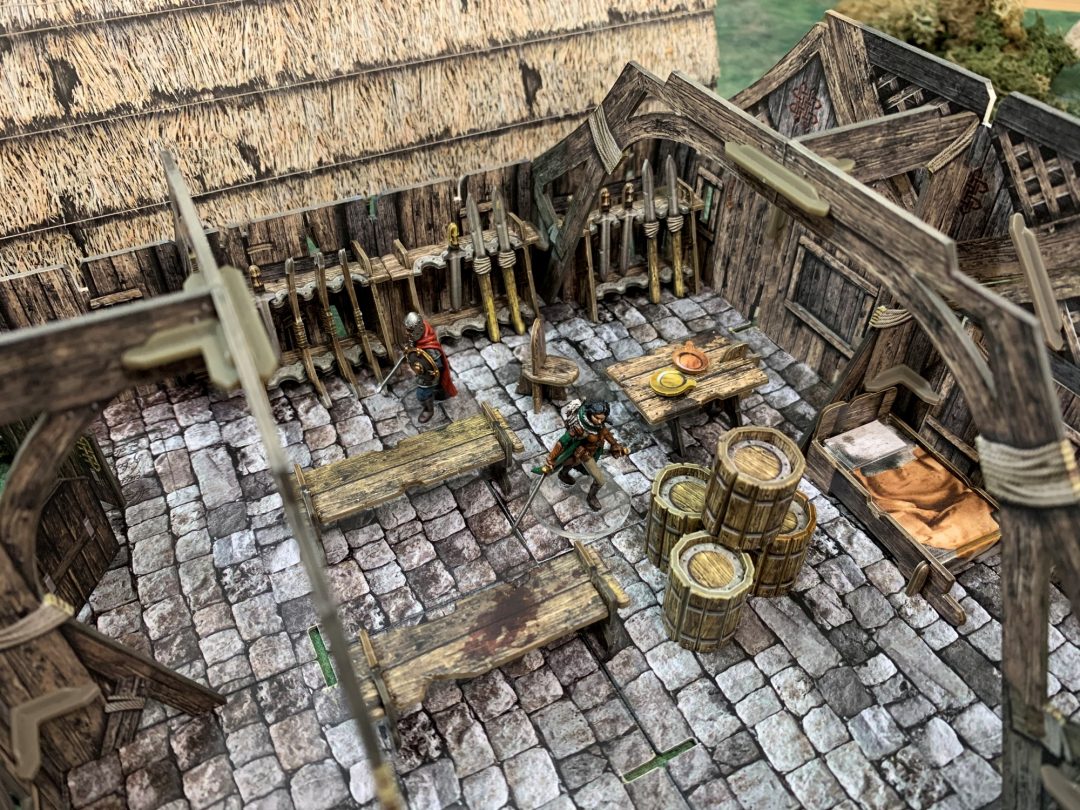 Two miniature figures stand inside a detailed wooden fantasy building with a stone floor, weapon racks, barrels, and furniture.