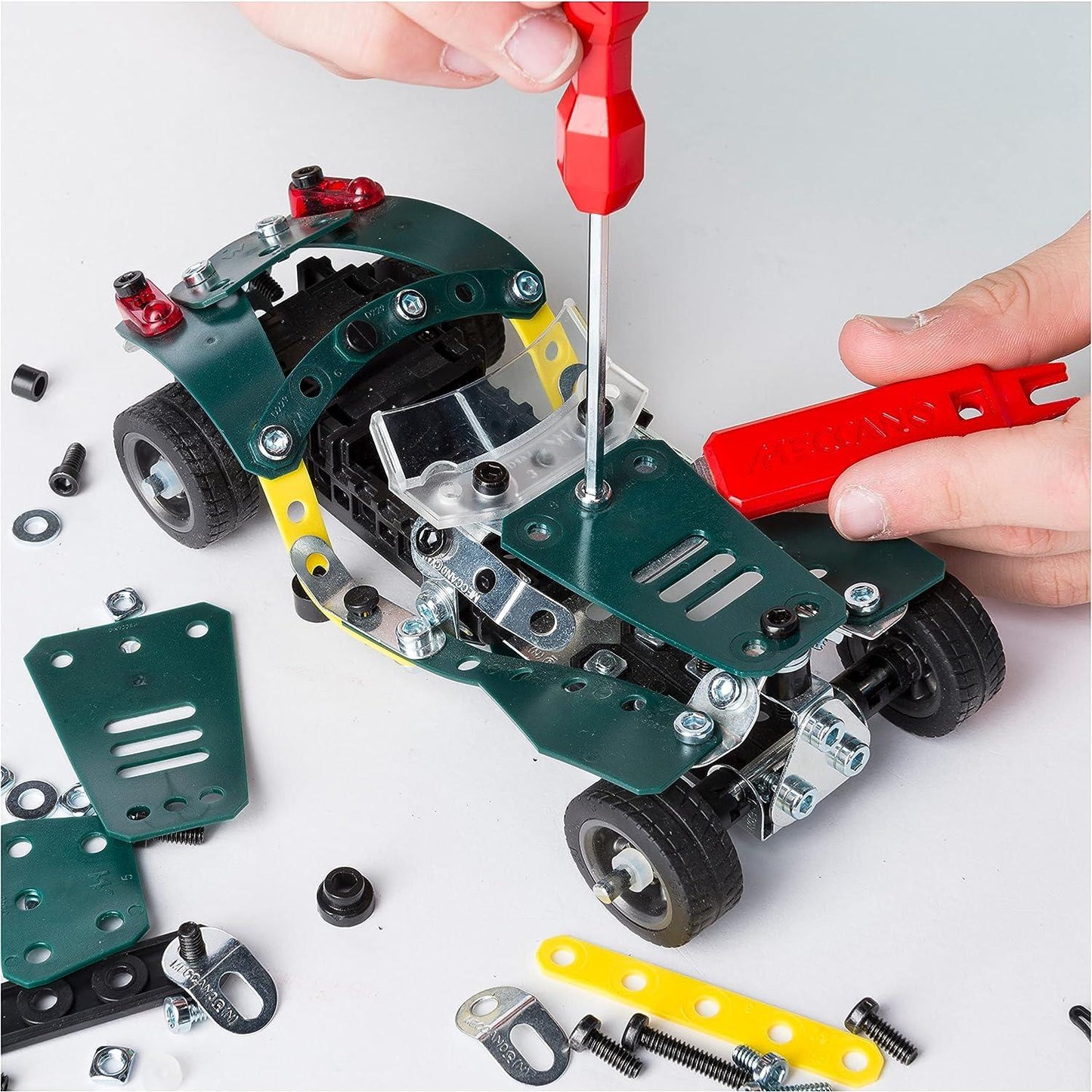 Hands assemble a green Meccano car using a red screwdriver and wrench with parts scattered around