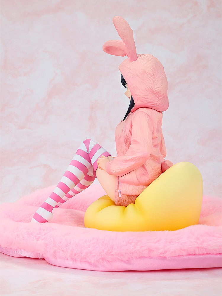 Anime figure in pink bunny onesie and striped socks, sitting on a yellow heart cushion on a pink fluffy surface