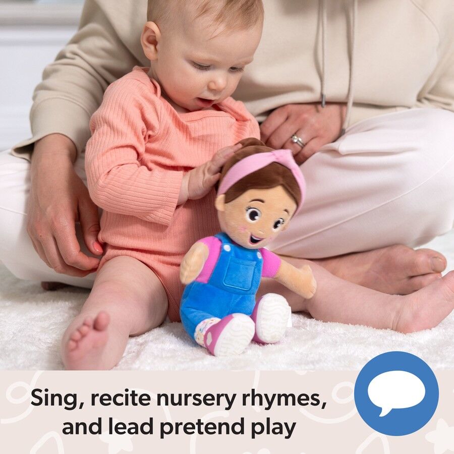 An infant in a peach outfit gently touches a plush girl doll wearing blue overalls and a pink headband, held by an adult