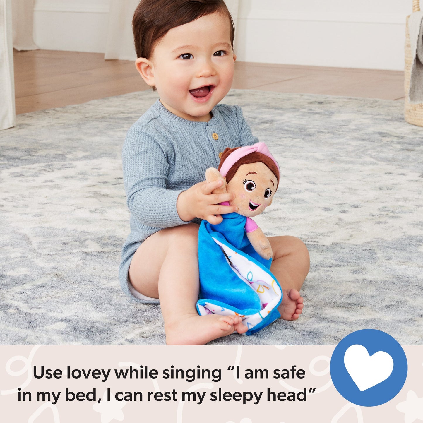 Happy baby with brown hair and light blue shirt, smiling and holding a soft lovey doll with a pink headband and blue blanket, sitting on a gray patterned rug