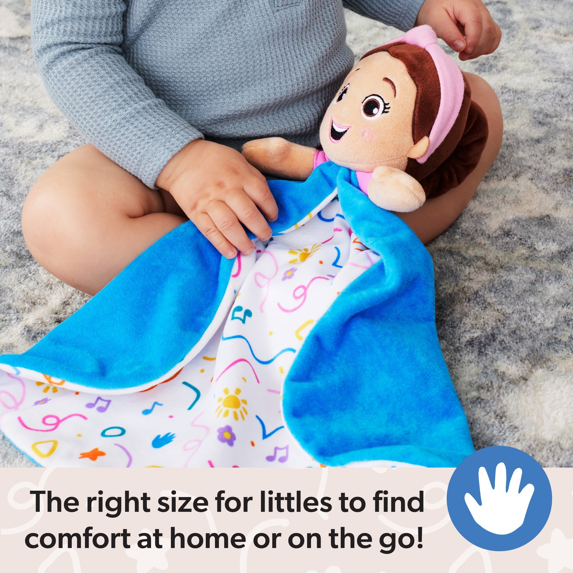 Toddler in a grey top holding a cheerful plush doll with a pink headband and a blue security blanket featuring colorful musical notes and suns, sitting on a soft rug