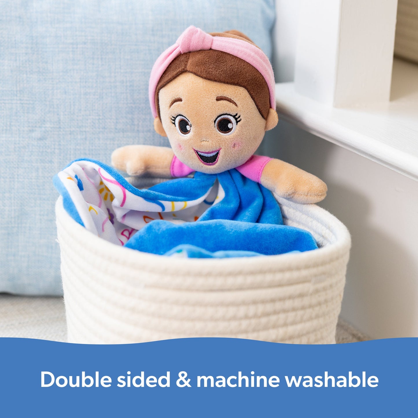 Smiling plush doll with pink headband and top, sits in a white woven basket with a blue blanket, features: double sided & machine washable