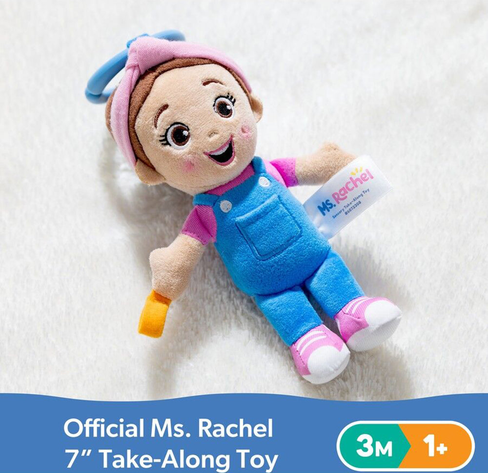 Smiling Ms. Rachel plush toy with pink headband, blue overalls, pink shirt, and pink shoes, lying on a white surface, a tag and blue clip are visible
