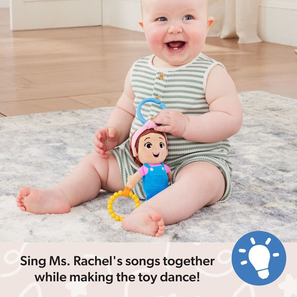 Happy baby in a striped romper holding a Ms. Rachel plush toy with a teether, sitting on a rug and smiling widely