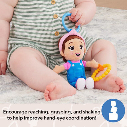 Baby in a green striped romper sits on a rug, holding a plush doll with a pink headband, blue overalls, and a yellow beaded rattle toy