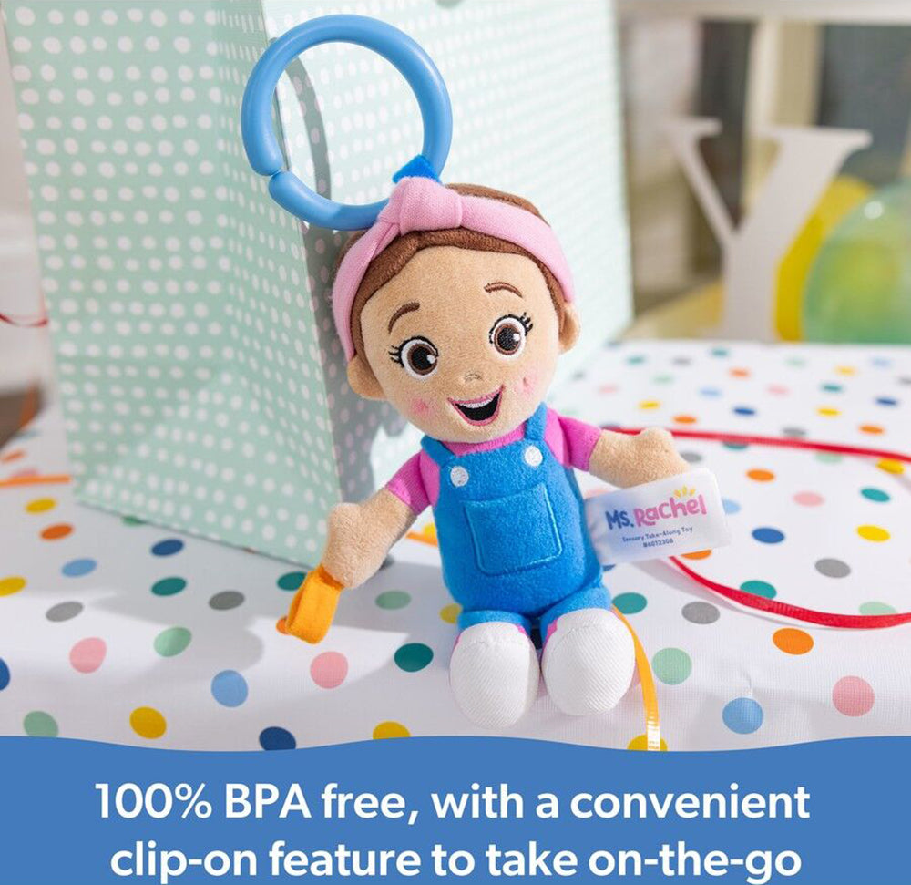 Ms. Rachel plush clip-on toy with pink headband, blue overalls, and orange teether, sitting on a polka dot table