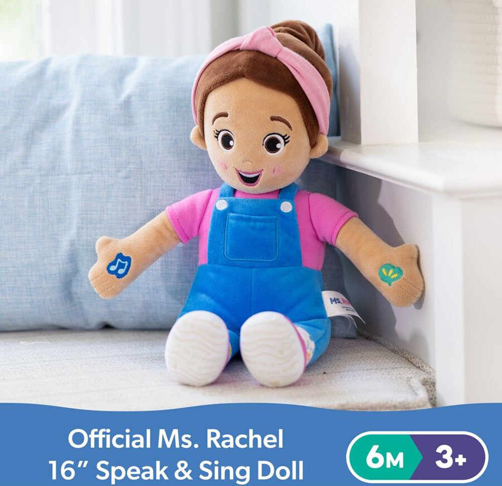 Official Ms. Rachel Speak & Sing plush doll: brown hair, pink headband, pink shirt, blue overalls, and musical note on left arm, sitting on a soft surface