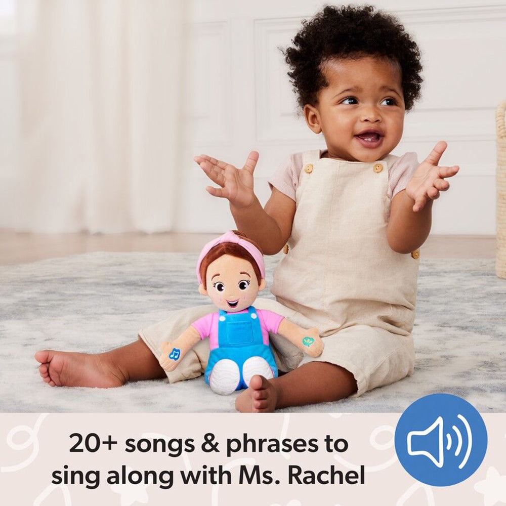 A happy baby with curly hair and a Ms. Rachel plush doll sit on a rug, the baby smiles, hands raised, wearing a beige romper